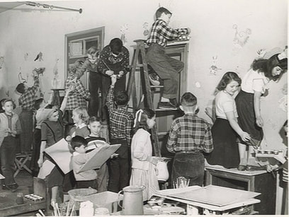 Black and white image of children standing on tables hanging art on the wall of a house, while other children stand behind them waiting with art in their arms.