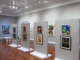 White gallery with 11 small works hanging on the wall at a distance and 5 pedestals with small sculpture resting on them in the center of the room.